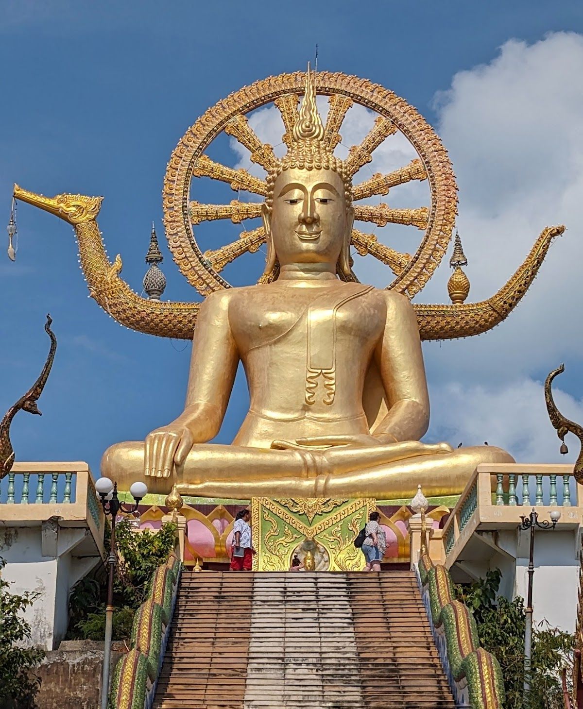 The golden Big Buddha statue at Wat Phra Yai, Koh Samui, viewed from below the steep naga-flanked staircase against a blue sky