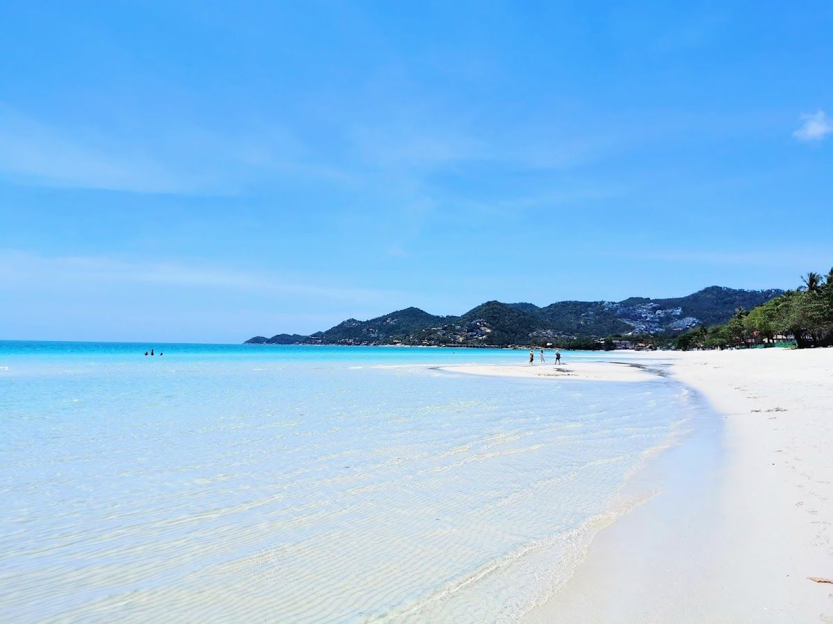 Chaweng Beach, Koh Samui — wide white sand with clear turquoise water and green hills in the background, a few people walking near the water's edge